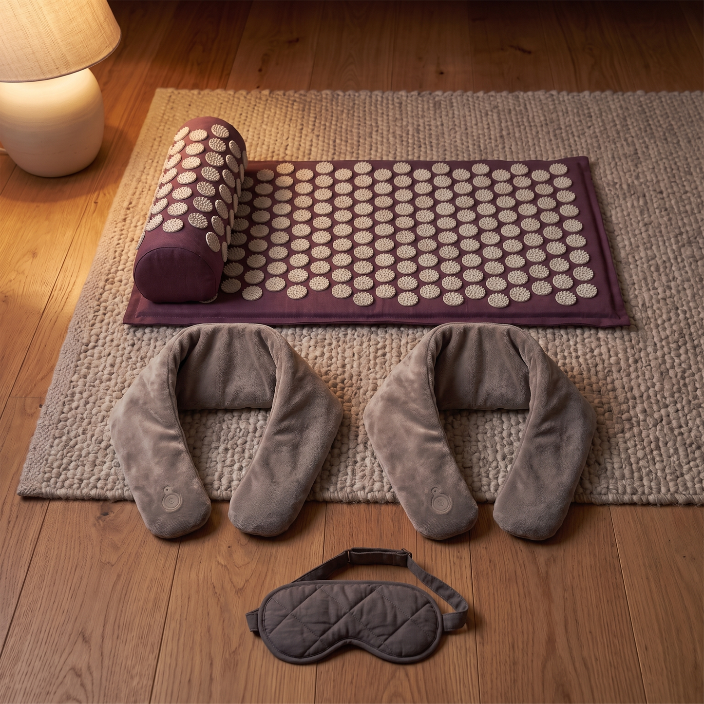 One plum acupressure mat and bolster, two grey heated neck wraps and a charcoal sleep mask laid out on a wool rug