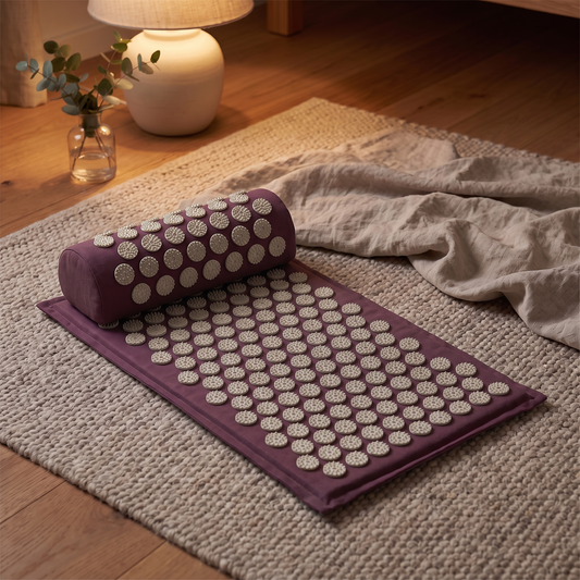 Plum acupressure mat and bolster pillow laid out on a wool rug in a warm lamplit living room