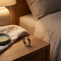 Stainless steel reset ring on an oiled-oak nightstand beside folded linen in warm lamplight