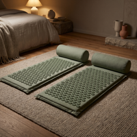 Two sage acupressure mats with their bolster pillows laid out side by side on a wool rug in warm lamplight