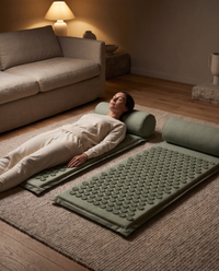 Woman lying back on one of two sage acupressure mats, the second mat and bolster waiting beside her