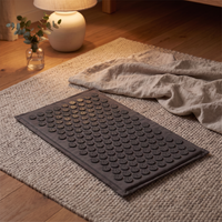 Charcoal acupressure mat laid out flat on a wool rug in a warm lamplit living room