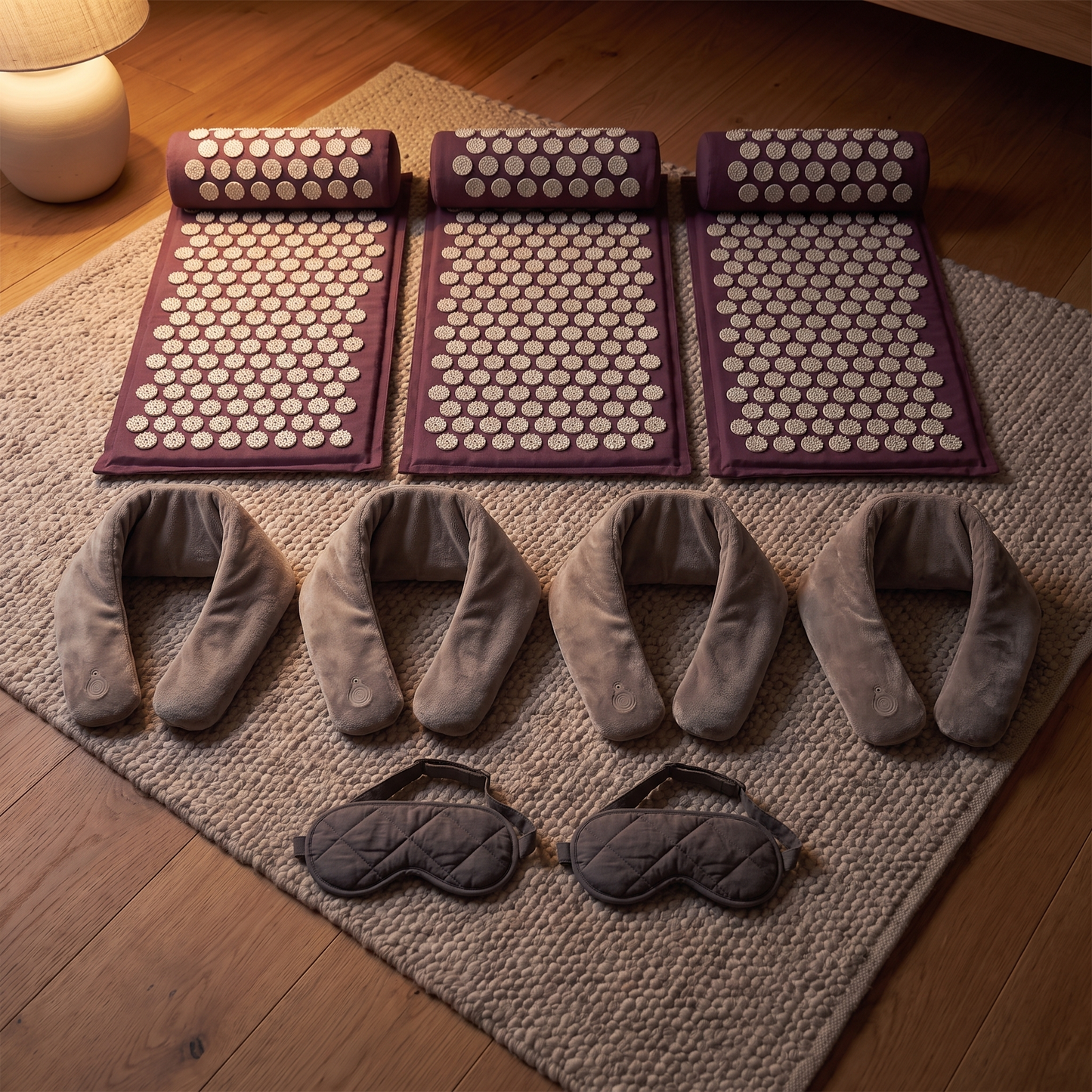 Three plum acupressure mats and bolsters, four grey heated neck wraps and two charcoal sleep masks laid out on a wool rug