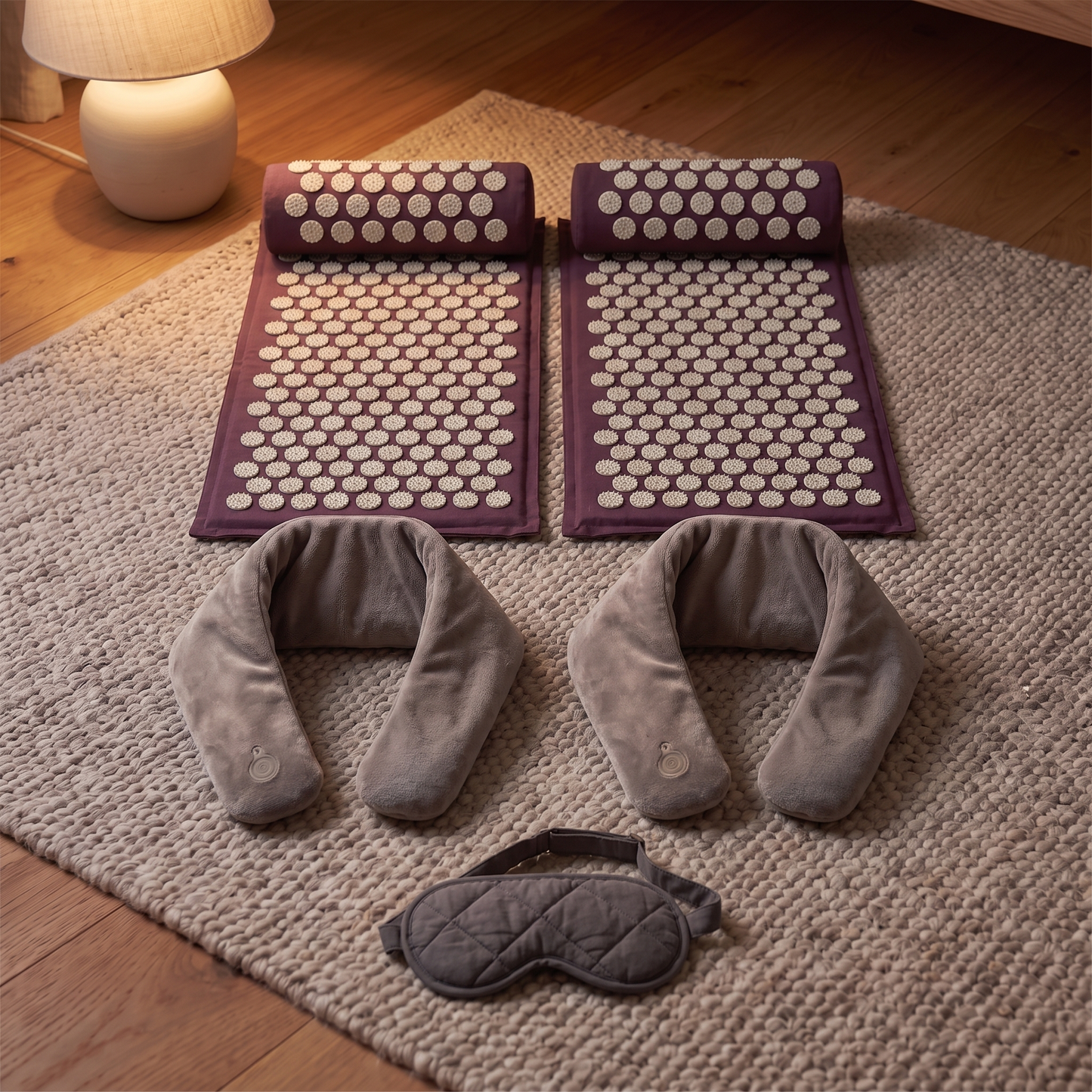 Two plum acupressure mats and bolsters, two grey heated neck wraps and a charcoal sleep mask laid out on a wool rug